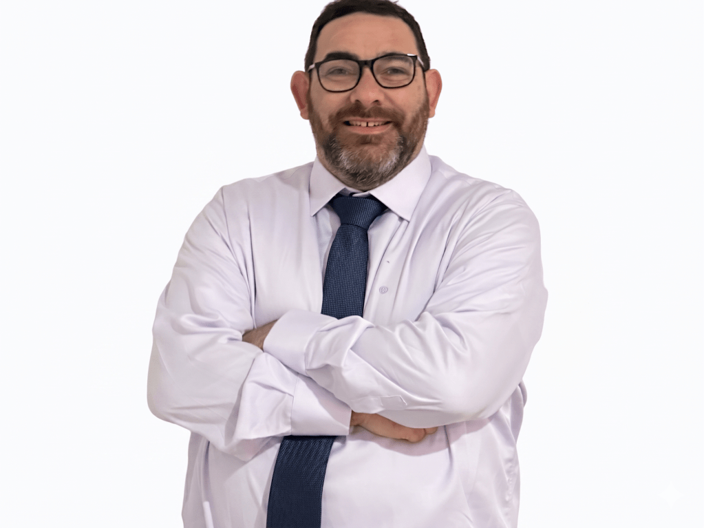 Retrato corporativo de plano medio de un hombre con gafas, barba y cabello oscuro, sonriendo con los brazos cruzados. Viste una camisa blanca de manga larga y una corbata azul oscuro, recortado sobre un fondo blanco puro sin sombras.